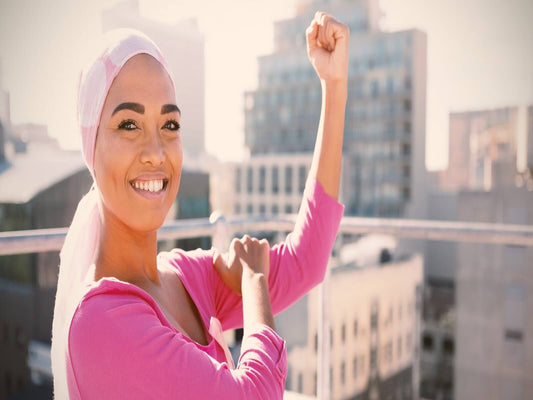 Woman standing on a rooftop, smiling and holding her arms up in celebration after beating cancer. She is wearing a pink shirt and a bandanna.