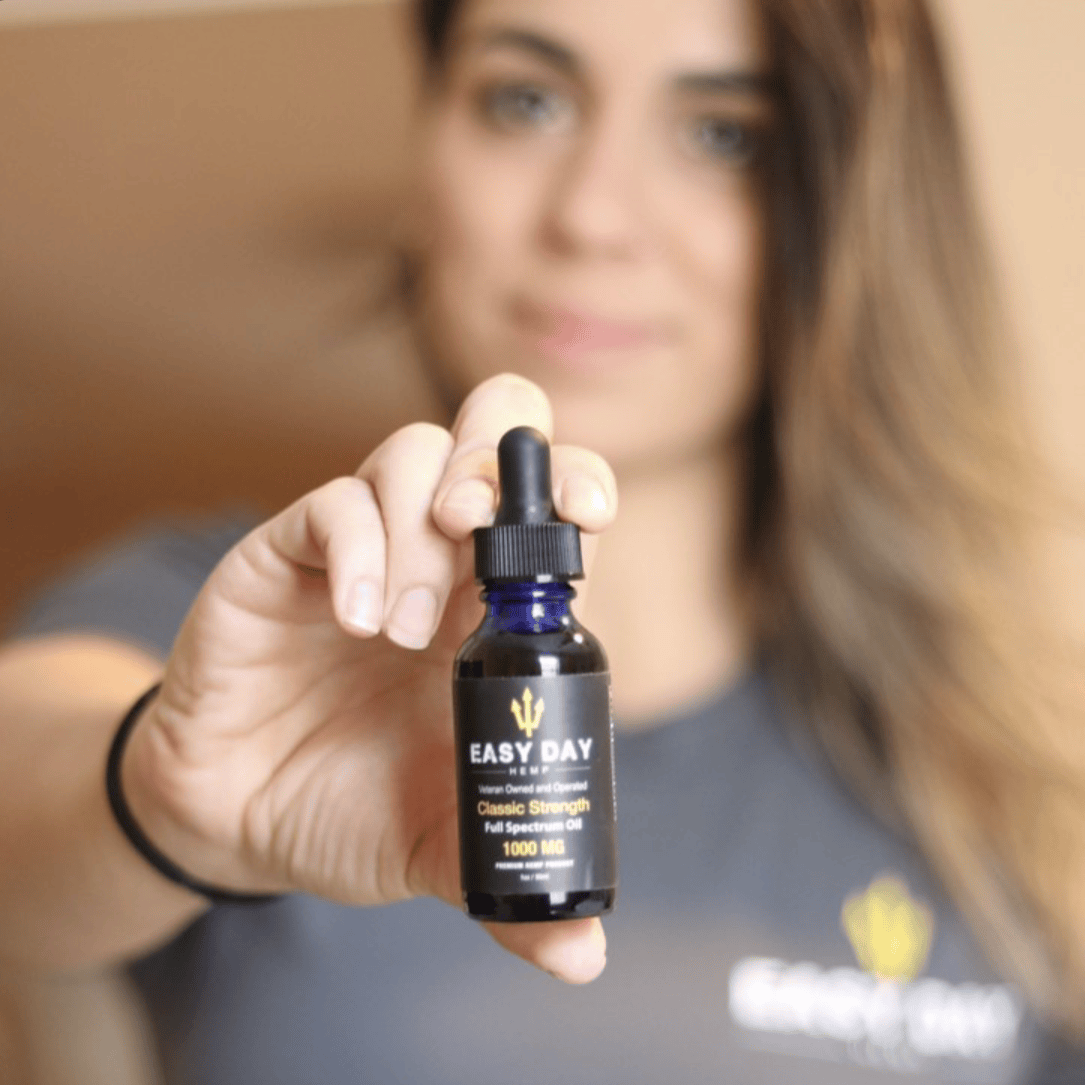 A woman with long, dark hair holding a bottle of Easy Day Hemp Classic strength 1000mg full spectrum CBD oil. The bottle is amber with a white label listing the ingredients and dosage instructions. The woman is looking at the bottle with a focused expression. This CBD oil is great for promoting overall wellness and may have potential benefits for anxiety, pain relief, and sleep
