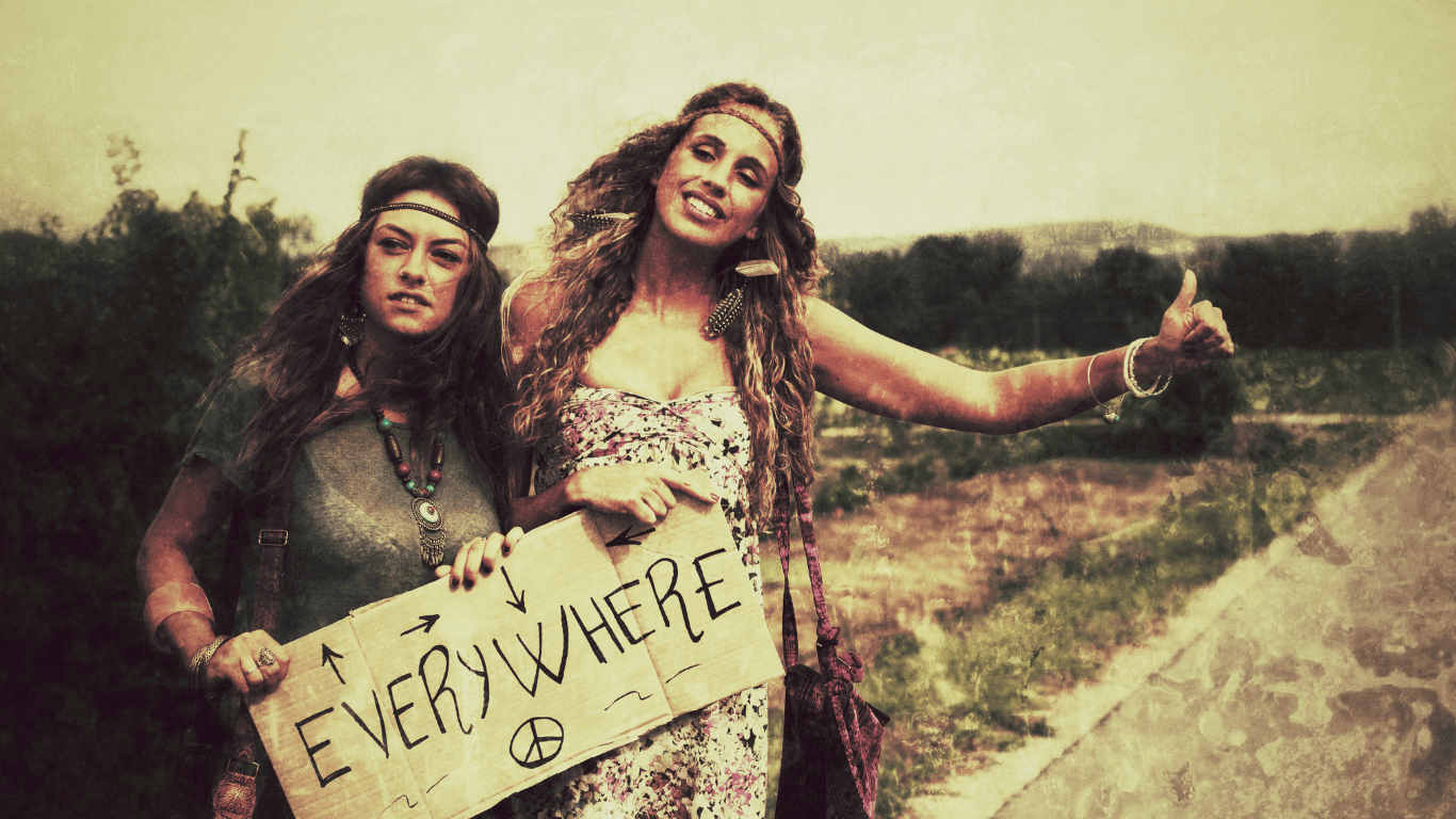A photograph of a hippie holding a cardboard sign that reads 'everywhere,' while hitchhiking. The hippie is dressed in traditional hippie fashion, with long hair and loose clothing.