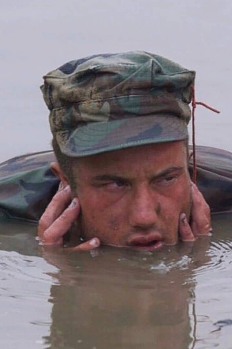A photograph of veteran Navy SEAL Jason Higgins going through the final day of Hell Week in basic underwater demolition SEAL training. Jason is wearing a camouflage hat and shirt, and appears tired with dirty water around his mouth. Hell Week is a grueling and challenging component of SEAL training, designed to test the physical and mental limits of candidates.