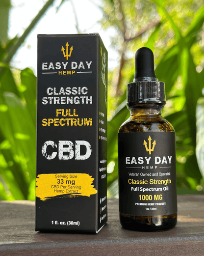 A brown bottle of Easy Day Hemp Classic strength 1000mg full spectrum CBD oil with a black label listing the ingredients and dosage instructions, sitting next to a box with a gold Easy Day logo and a jungle background. This CBD oil is great for promoting overall wellness and may have potential benefits for anxiety, pain relief, and sleep.