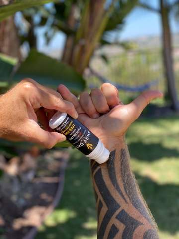 A veteran Navy SEAL named Jason Higgins holding the Elite CBD Pain Roll-On in his left hand and applying it to his right arm, which is tattooed with black tribal artwork. His hand is in the "tuanis" symbol, which is common among surfers. This CBD product may have potential benefits for pain relief and overall wellness.